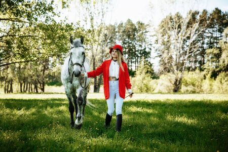 Beautiful young girl with blond hair in a red jacket with a fringe , wearing red hat, smiling and stroking her horse in summer field. equestrian sport conceptの写真素材