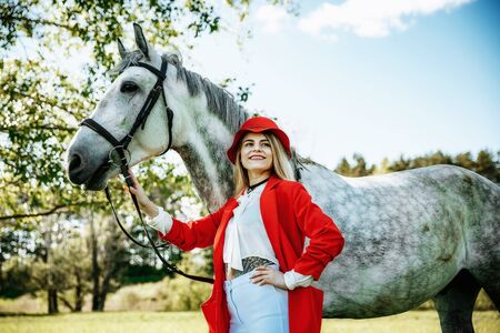 Beautiful young girl with blond hair in a red jacket with a fringe , wearing red hat, smiling and stroking her horse in summer field. equestrian sport conceptの写真素材