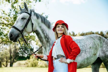 Beautiful young girl with blond hair in a red jacket with a fringe , wearing red hat, smiling and stroking her horse in summer field. equestrian sport conceptの写真素材