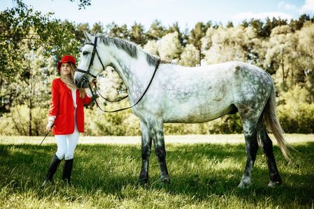 beauty blondie woman in red jacket and red hat with horse in the field, effect of toning. Equestrian sport concept. Fashion photoshootの写真素材