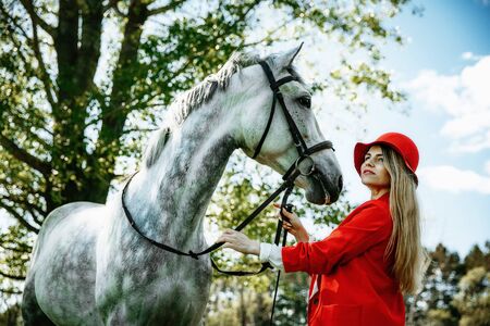 beauty blondie woman in red jacket and red hat with horse in the field, effect of toning. Equestrian sport concept. Fashion photoshootの写真素材