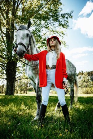 beauty blondie woman in red jacket and red hat with horse in the field, effect of toning. Equestrian sport concept. Fashion photoshootの写真素材