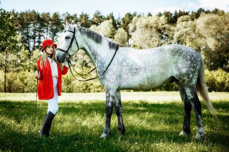beauty blondie woman in red jacket and red hat with horse in the field, effect of toning. Equestrian sport concept. Fashion photoshootの写真素材