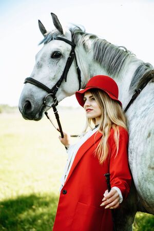 Beautiful young girl with blond hair in a red jacket with a fringe , wearing red hat, smiling and stroking her horse in summer field. equestrian sport conceptの写真素材