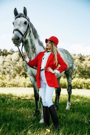Beautiful young girl with blond hair in a red jacket with a fringe , wearing red hat, smiling and stroking her horse in summer field. equestrian sport conceptの写真素材