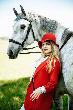 beauty blondie woman in red jacket and red hat with horse in the field, effect of toning. Equestrian sport concept. Fashion photoshootの写真素材