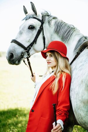 beauty blondie woman in red jacket and red hat with horse in the field, effect of toning. Equestrian sport concept. Fashion photoshootの写真素材