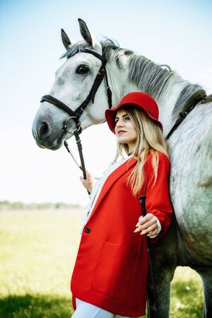 Beautiful young girl with blond hair in a red jacket with a fringe , wearing red hat, smiling and stroking her horse in summer field. equestrian sport conceptの写真素材