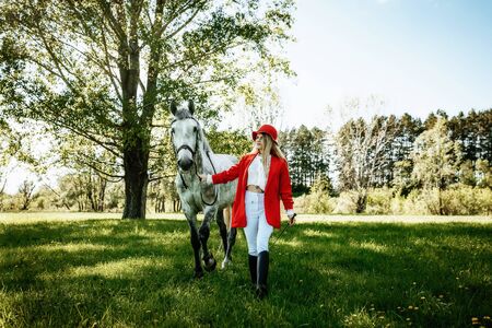 Beautiful young girl with blond hair in a red jacket with a fringe , wearing red hat, smiling and stroking her horse in summer field. equestrian sport conceptの写真素材