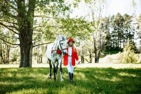 Beautiful young girl with blond hair in a red jacket with a fringe , wearing red hat, smiling and stroking her horse in summer field. equestrian sport conceptの写真素材