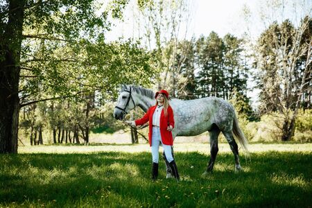 Beautiful young girl with blond hair in a red jacket with a fringe , wearing red hat, smiling and stroking her horse in summer field. equestrian sport conceptの写真素材