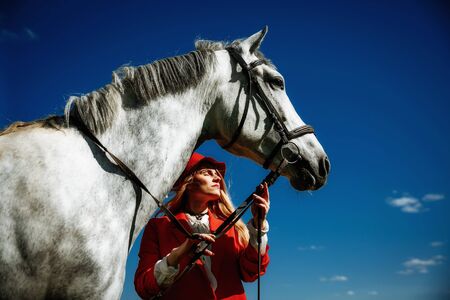 Beautiful young girl with blond hair in a red jacket with a fringe , wearing red hat, smiling and stroking her horse in summer field. equestrian sport conceptの写真素材