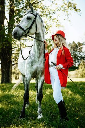 Beautiful young girl with blond hair in a red jacket with a fringe , wearing red hat, smiling and stroking her horse in summer field. equestrian sport conceptの写真素材