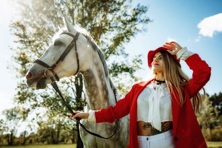 Beautiful young girl with blond hair in a red jacket with a fringe , wearing red hat, smiling and stroking her horse in summer field. equestrian sport conceptの写真素材