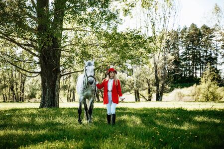 Beautiful young girl with blond hair in a red jacket with a fringe , wearing red hat, smiling and stroking her horse in summer field. equestrian sport conceptの写真素材