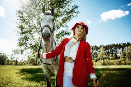 Beautiful young girl with blond hair in a red jacket with a fringe , wearing red hat, smiling and stroking her horse in summer field. equestrian sport conceptの写真素材