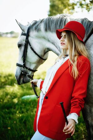 beauty blondie woman in red jacket and red hat with horse in the field, effect of toning. Equestrian sport concept. Fashion photoshootの写真素材