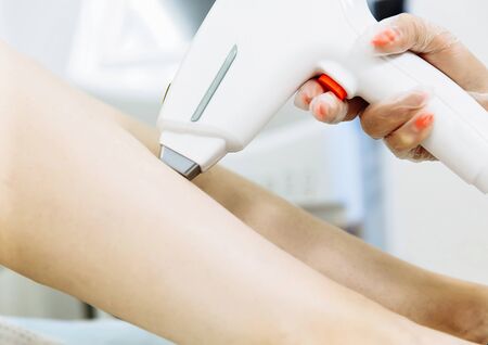 Close up of beautician removing hair on lady leg with laser epilation machine. Girl sitting on comfortable daybedの写真素材
