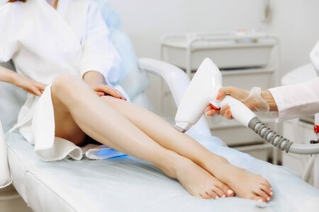 Close up of beautician removing hair on lady leg with laser epilation machine. Girl sitting on comfortable daybedの写真素材