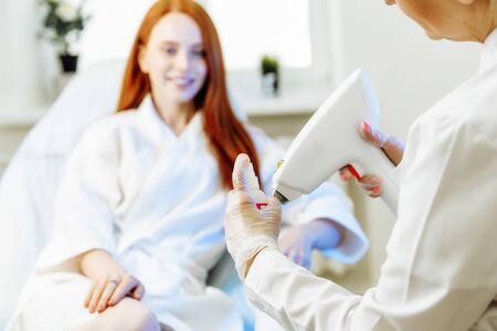 The doctor is preparing a laser hair removal machine for work. Against the background is the woman who will be given the procedure. A man and a doctor are in a modern beauty salon.の写真素材