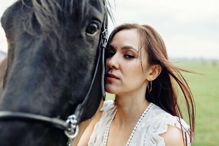 Adorable woman in white dress standing with black horse in fieldの写真素材