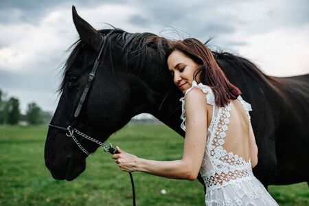 Adorable woman in white dress standing with black horse in fieldの写真素材