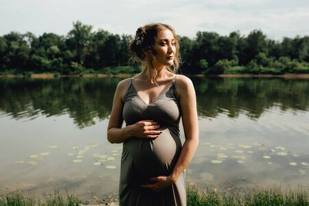 Beautiful pregnant woman walks in the green park along the riverの写真素材
