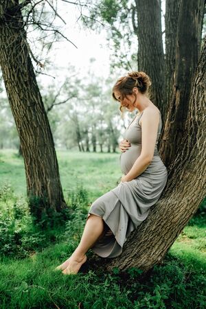Pregnant woman posing in the park . Portrait of a happy and proud pregnant woman looking at her bellyの写真素材