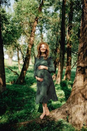 Smiling pretty pregnant woman touching around the abdomen against greenery background. Wearing green dress and summer hatの写真素材