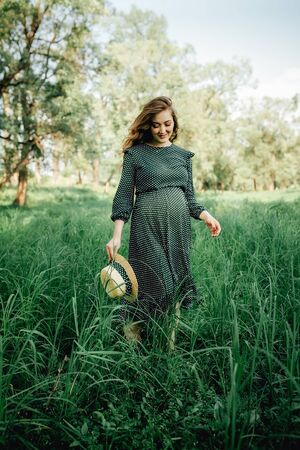 Smiling pretty pregnant woman touching around the abdomen against greenery background. Wearing green dress and summer hatの写真素材