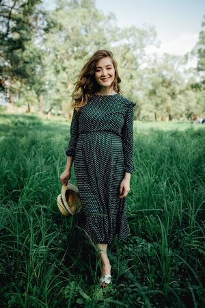 Smiling pretty pregnant woman touching around the abdomen against greenery background. Wearing green dress and summer hatの写真素材