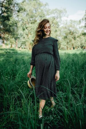 Smiling pretty pregnant woman touching around the abdomen against greenery background. Wearing green dress and summer hatの写真素材