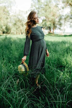 Smiling pretty pregnant woman touching around the abdomen against greenery background. Wearing green dress and summer hatの写真素材