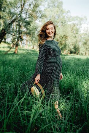 Smiling pretty pregnant woman touching around the abdomen against greenery background. Wearing green dress and summer hatの写真素材