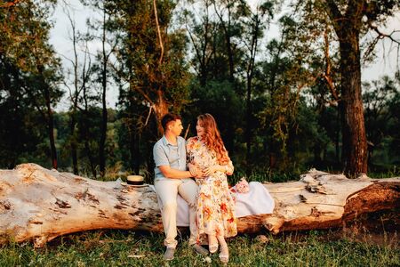 Stylish pregnant couple holding hands on belly in sunny light in summer park. Happy young parents, mom and dad, hugging baby bump and smiling, enjoying beautiful moment at sunset among grassの写真素材