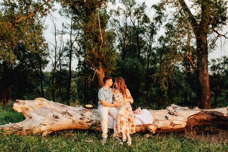 Stylish pregnant couple holding hands on belly in sunny light in summer park. Happy young parents, mom and dad, hugging baby bump and smiling, enjoying beautiful moment at sunset among grassの写真素材
