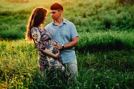 Happy young parents, mom and dad, hugging baby bump and smiling, enjoying beautiful moment at sunset among grass. Stylish pregnant couple holding hands on belly in sunny light in summer park.の写真素材