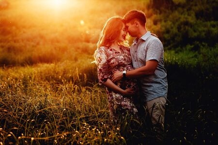 Happy young parents, mom and dad, hugging baby bump and smiling, enjoying beautiful moment at sunset among grass. Stylish pregnant couple holding hands on belly in sunny light in summer park.の写真素材