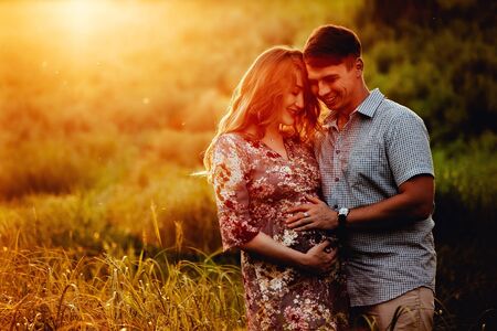 Happy young parents, mom and dad, hugging baby bump and smiling, enjoying beautiful moment at sunset among grass. Stylish pregnant couple holding hands on belly in sunny light in summer park.の写真素材