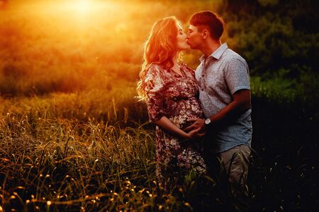 Happy young parents, mom and dad, hugging baby bump and smiling, enjoying beautiful moment at sunset among grass. Stylish pregnant couple holding hands on belly in sunny light in summer park.の写真素材