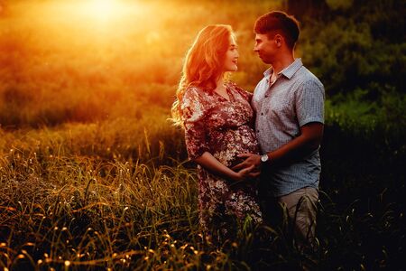 Young happy beautiful couple in love walking together on grass and trees park landscape on sunset in summer with man touching pregnant belly of his woman. Family conceptの写真素材