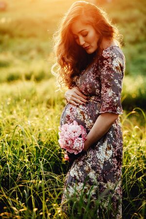 Pregnant woman feeling happy with new life at summer field while take care of her child. The young expecting mother holding baby in pregnant belly. Maternity prenatal care and woman pregnancy concept.の写真素材