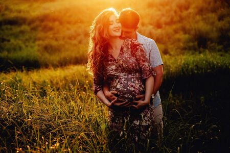 Young happy beautiful couple in love walking together on grass and trees park landscape on sunset in summer with man touching pregnant belly of his woman. Family conceptの写真素材