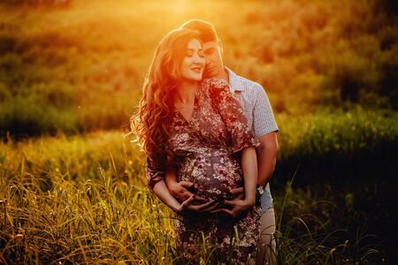 Happy young parents, mom and dad, hugging baby bump and smiling, enjoying beautiful moment at sunset among grass. Stylish pregnant couple holding hands on belly in sunny light in summer park.の写真素材