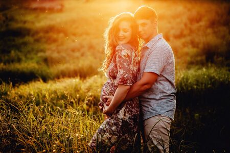 Happy young parents, mom and dad, hugging baby bump and smiling, enjoying beautiful moment at sunset among grass. Stylish pregnant couple holding hands on belly in sunny light in summer park.の写真素材
