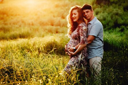 Happy young parents, mom and dad, hugging baby bump and smiling, enjoying beautiful moment at sunset among grass. Stylish pregnant couple holding hands on belly in sunny light in summer park.の写真素材