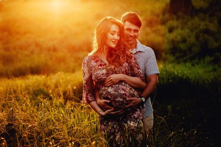 Young happy beautiful couple in love walking together on grass and trees park landscape on sunset in summer with man touching pregnant belly of his woman. Family conceptの写真素材