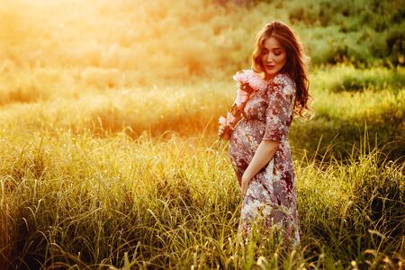 Pregnant woman feeling happy with new life at summer field while take care of her child. The young expecting mother holding baby in pregnant belly. Maternity prenatal care and woman pregnancy concept.の写真素材
