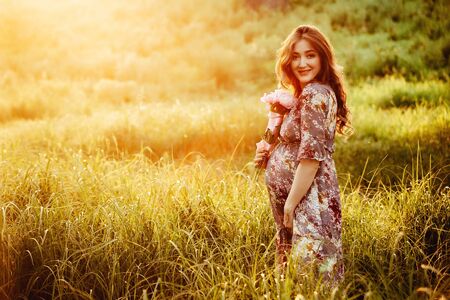 Pregnant woman feeling happy with new life at summer field while take care of her child. The young expecting mother holding baby in pregnant belly. Maternity prenatal care and woman pregnancy concept.の写真素材