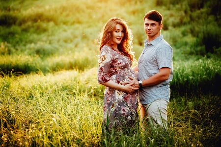 Happy young parents, mom and dad, hugging baby bump and smiling, enjoying beautiful moment at sunset among grass. Stylish pregnant couple holding hands on belly in sunny light in summer park.の写真素材